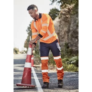A man wearing high visibility orange and navy work pants adjusts a traffic cone on a rural road.