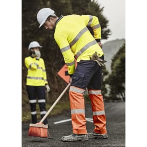 Hi-vis yellow and orange work pants with reflective tape, worn by a worker sweeping the road. A colleague is in the background.