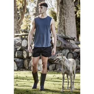 A man in a sleeveless top and black rugby shorts walks beside a greyhound in a natural setting.