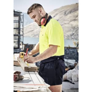 A man in a high-visibility yellow shirt and navy rugby shorts measures a wooden plank at a construction site.