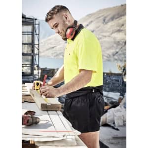 A man in a high-visibility yellow shirt and black sports shorts measures a piece of wood at a construction site.