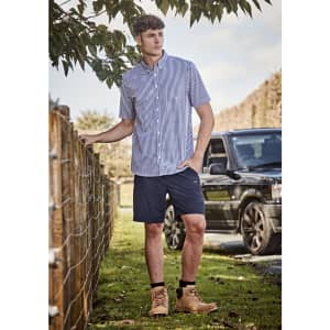 A man wearing a short-sleeve checked shirt and navy lightweight outdoor shorts, standing by a fence.