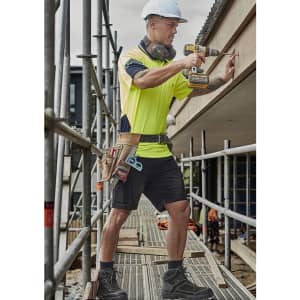 A man in a high-visibility shirt and cargo shorts uses a drill on a construction site, wearing safety gear and a tool belt.
