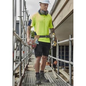 A man wearing a high-visibility yellow polo shirt and black cargo shorts, carrying tools on a construction site.