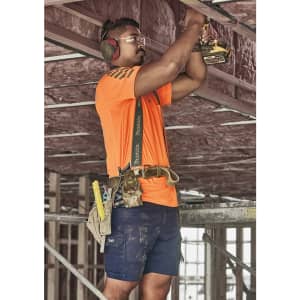 A man wearing an orange shirt and navy blue cargo shorts works on a construction site, using a power drill.