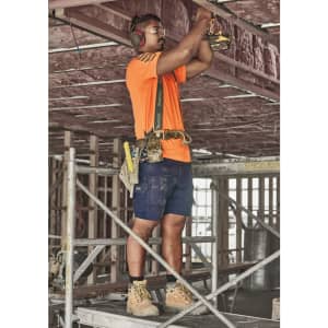 A worker in an orange shirt and navy cargo shorts uses a power tool on a construction site.