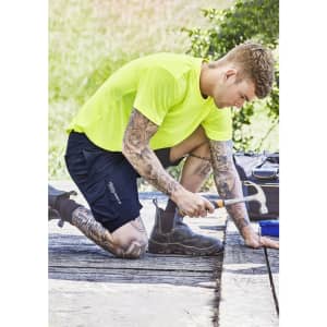 A person kneeling outdoors in bright yellow clothing and dark blue sports shorts, using a saw on a wooden surface.