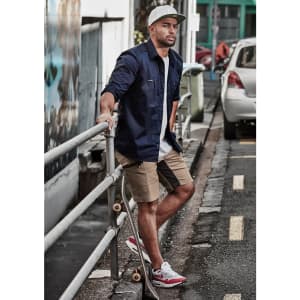 A man wearing a navy shirt and beige stretch shorts stands by a railing, with a skateboard beside him.