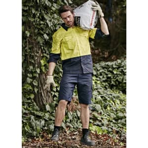 Men's rugged cooling vented shorts in navy, paired with a yellow work shirt, set against a lush green background.