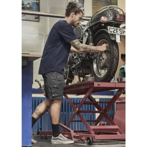 A man in navy shorts works on a motorcycle in a garage, featuring a logo on the shorts.