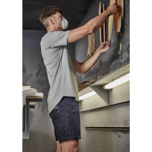 A man wearing a light grey shirt and dark shorts is organising materials on a wall in a workshop.