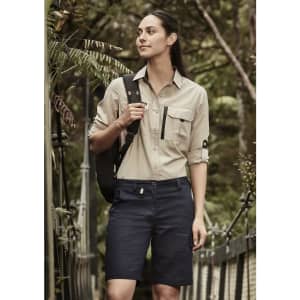 A woman in a light beige shirt and navy shorts stands outdoors, with a backpack over her shoulder. The shorts feature a logo.