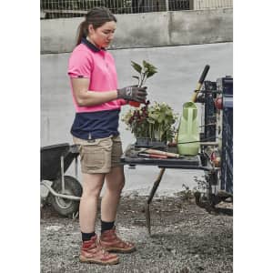 A woman in a pink and navy polo shirt wears beige cargo shorts, holding plants beside gardening tools and a wheelbarrow.