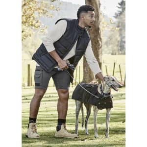 A man wearing a black vest and grey shorts stands with a leashed greyhound in a grassy park.