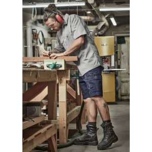 A man in a grey shirt and navy shorts, wearing safety gear, works at a woodworking bench in a workshop.