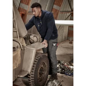 A man in a navy 1/4 zip polar fleece works on an engine in a workshop, wearing work pants with pockets.