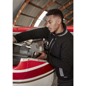 A man in a black fleece jacket works on an aircraft engine inside a hangar. The jacket features a logo.