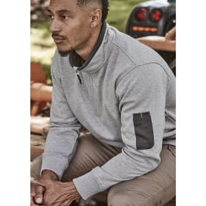 A man wearing a grey unisex 1/4 zip brushed fleece pullover with a logo on the sleeve, seated outdoors.