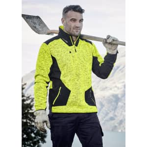 A man holds a shovel while wearing a bright yellow and black reinforced knit 1/2 zip pullover, set against a mountainous backdrop.