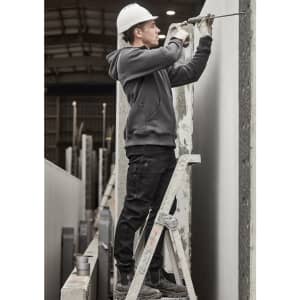 A person in a grey zip front multi-pocket hoodie and black pants using a drill while standing on a ladder.