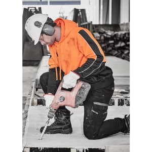 A man in an orange hi-vis pullover hoodie using a drill on a construction site, wearing a safety helmet and ear protection.