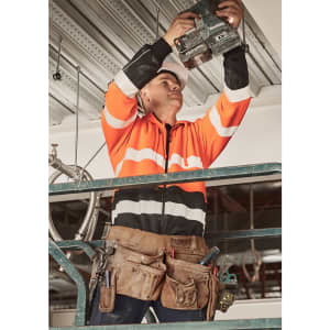 A man in an orange and black hi-vis hoodie uses a tool while standing on a scaffold, wearing a tool belt.