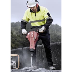 A worker in a fluorescent yellow and black hooped pullover operates a demolition tool at a construction site.