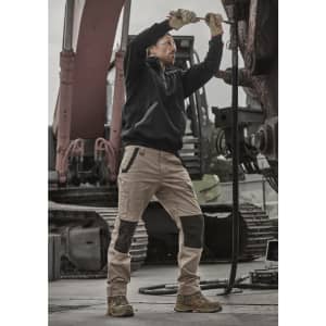 A man wears a black pullover while working with machinery, paired with beige and brown pants, in an industrial setting.