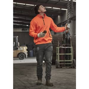 A unisex water-resistant 1/4 zip pullover in bright orange, worn with dark grey trousers, set in an industrial environment.