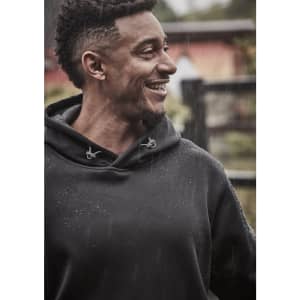 A man wearing a black unisex water-resistant hoodie, smiling as rain falls around him. The hoodie features a logo.