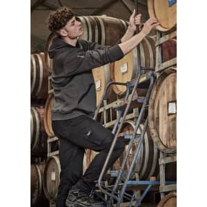 A person in a dark grey unisex water-resistant hoodie climbing a ladder among wine barrels. The hoodie has a logo.