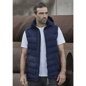 A man wearing a navy blue unisex hooded puffer vest over a white t-shirt, standing against a backdrop of pipes.