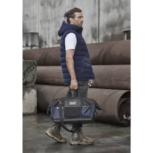 A man wearing a navy hooded puffer vest holds a black and navy duffle bag, standing against industrial backdrops.