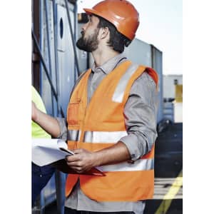 A man wearing an orange hi-vis zip vest with reflective stripes and a hard hat, holding documents at a construction site.