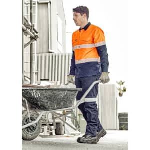 A man in an orange and navy utility pant pushes a wheelbarrow at a construction site. The pants have reflective detailing and a logo.