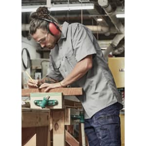 A lightweight grey short sleeve work shirt worn by a man using a pencil on wood in a workshop. The shirt has a logo.