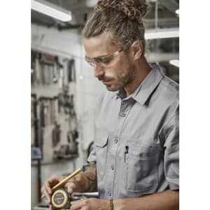 A man wearing a grey short sleeve work shirt, safety glasses, examining a measuring tape in a workshop setting.