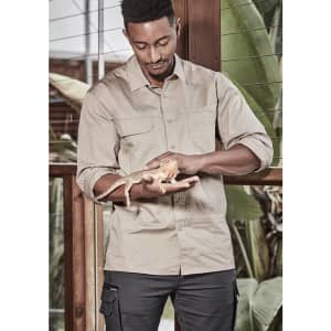A man wearing a beige long sleeve work shirt holds a lizard in a tropical setting. The shirt features a logo.