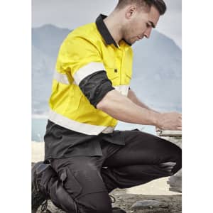 A man kneeling outdoors in a yellow and black long sleeve work shirt with reflective tape, engaged in construction work.