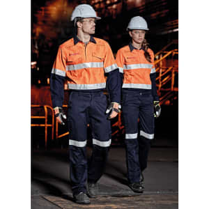 Two individuals wearing orange and navy safety shirts and pants, equipped with hard hats and gloves, in an industrial setting.