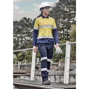 A woman in a yellow and navy lightweight ripstop taped shirt walks on a wooden path, wearing a helmet and holding gloves.