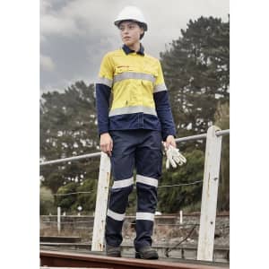 A woman in a yellow and navy work shirt with reflective stripes, wearing a hard hat and gloves, stands outdoors.