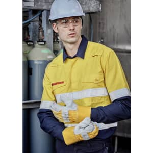 A man in a yellow and navy work shirt with reflective stripes, wearing protective goggles and gloves, standing near gas cylinders.