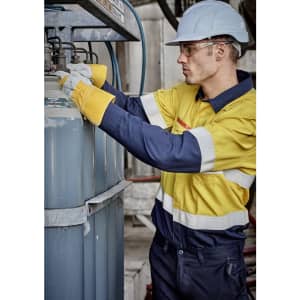 A worker in a yellow and navy spliced shirt adjusts equipment, wearing a hard hat and gloves in an industrial setting.