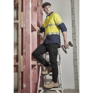 A man in a yellow and navy long sleeve shirt stands on a ladder, holding power tools, next to timber framing.