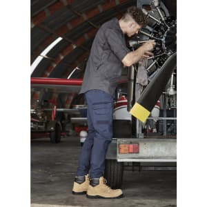 A man in a dark grey shirt and navy pants works on a machine, wearing tan boots in a hangar setting.
