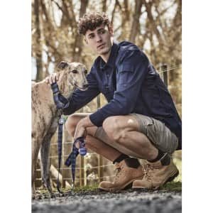 A man in a navy long sleeve button-up shirt kneels next to a greyhound on a leash. The background features trees.