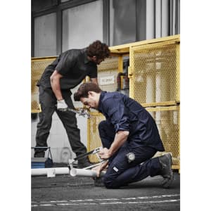 Two men in dark work shirts and pants engage in plumbing tasks outdoors near a yellow fence.