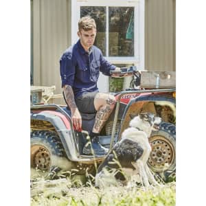 A man in a navy long sleeve button-up shirt sits on an all-terrain vehicle beside a dog. The setting is outdoor with grass.