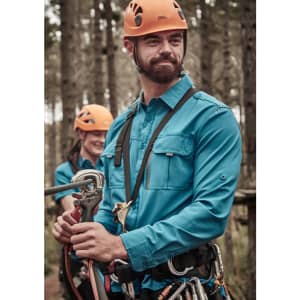 A blue long sleeve button-up shirt worn by a man, in a forest setting, with climbing gear and a helmet.
